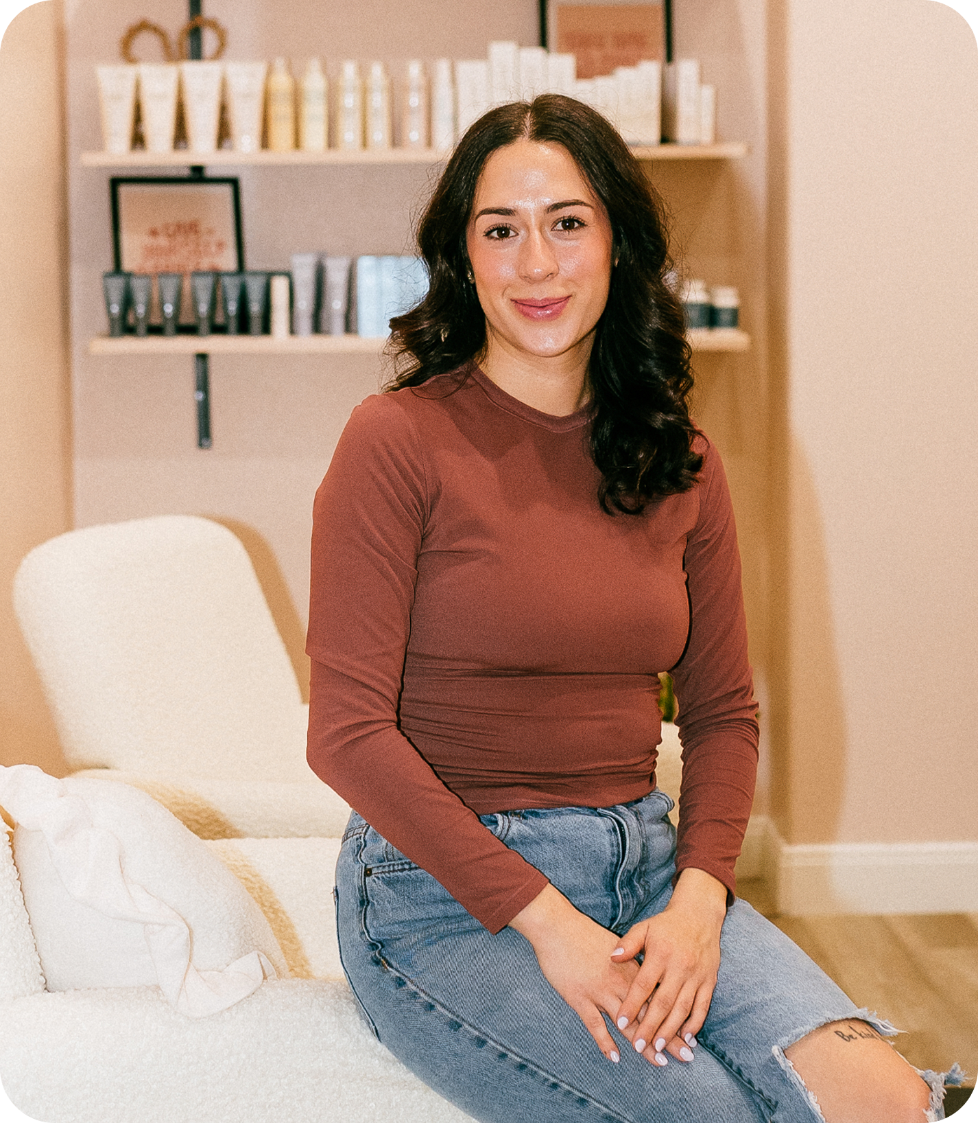 Women in a tan top and jeans smiling and sitting on a bed