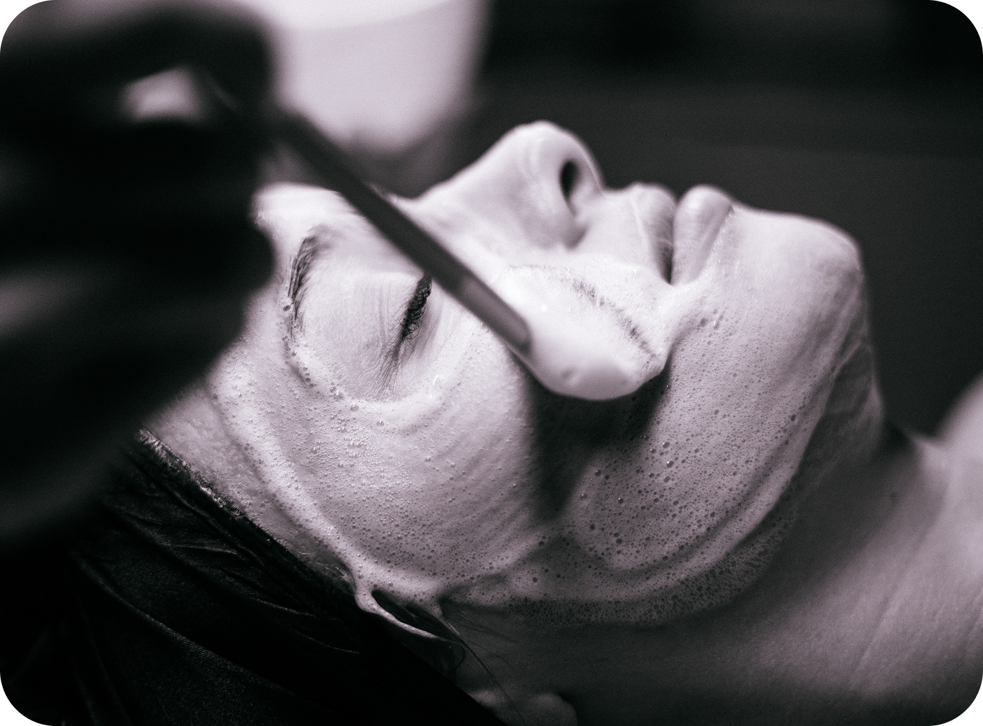 Women with her eyes closed laying down during a facial and a fan brush with cleanser on it going over her face.
