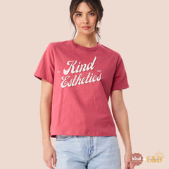 Person wearing a pink t-shirt with 'Kind Esthetics' text on a beige background